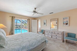 Une chambre à coucher comprenant un lit, une commode, une chaise, un ventilateur de plafond, une fenêtre avec des rideaux et un tableau accroché au mur.