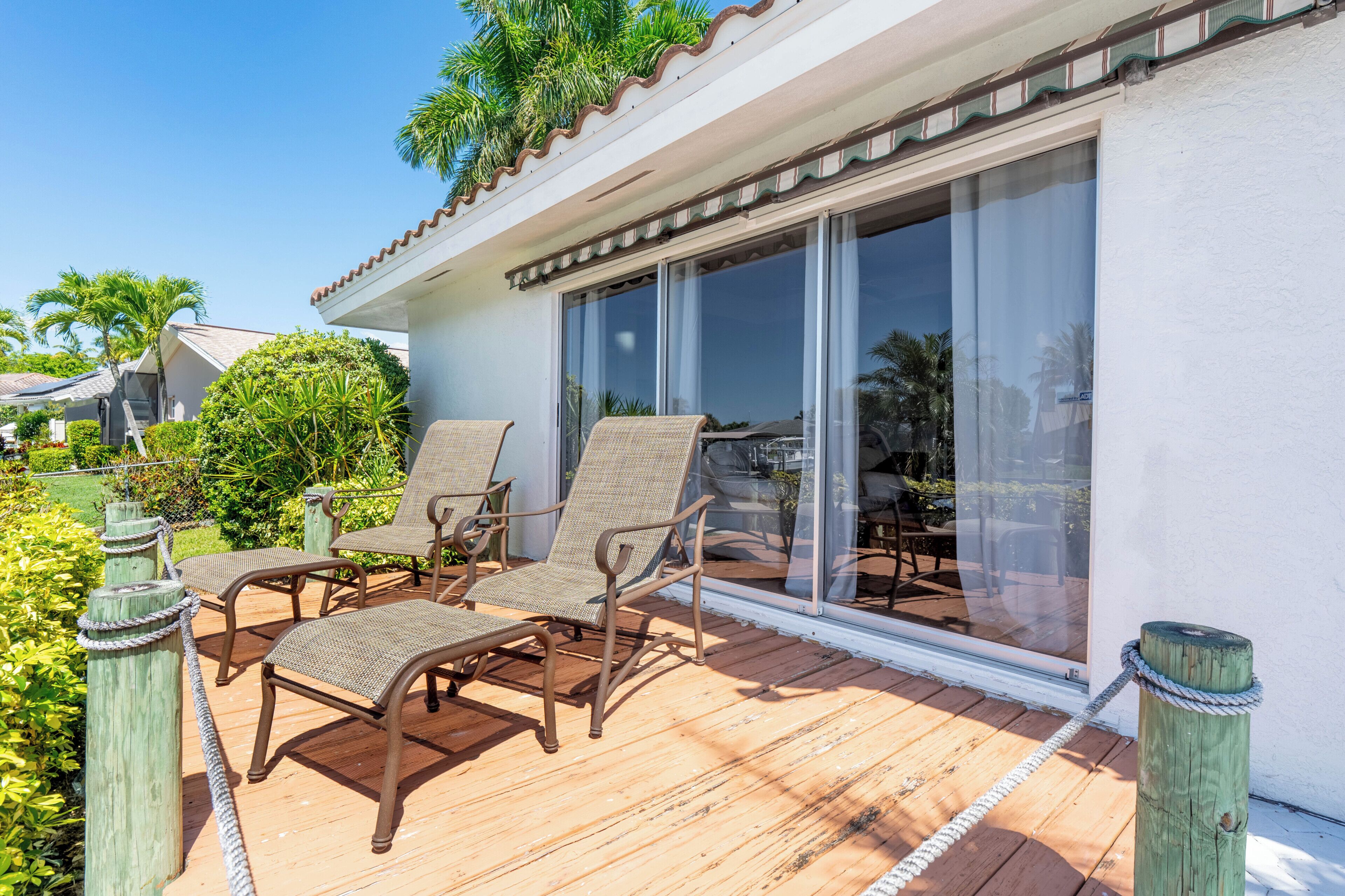 Une terrasse en bois avec deux fauteuils en osier, une porte vitrée et un bâtiment blanc doté d’un toit en tuiles.