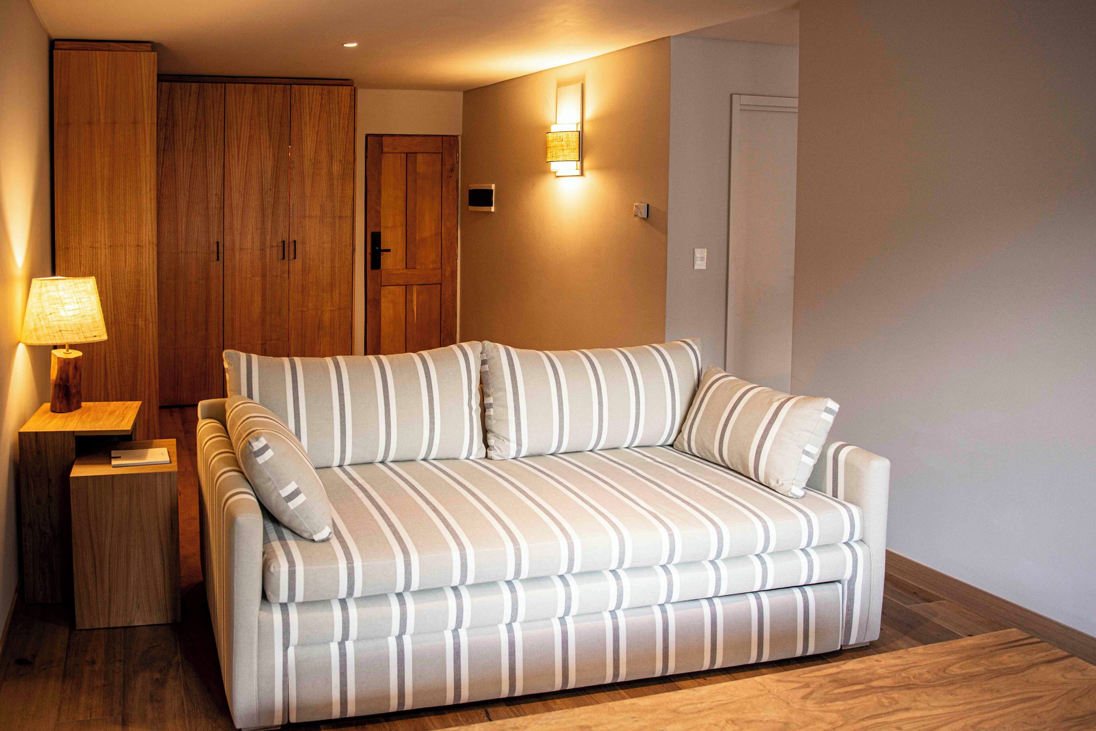 A room with a striped sofa, a wooden wardrobe, a bedside table with a lamp, and a wall-mounted light fixture.
