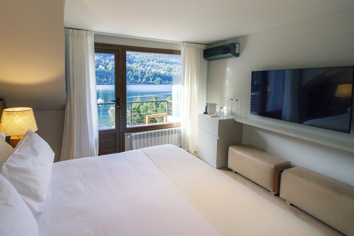 Ein Hotelzimmer mit einem großen Bett, einem Fernseher, einem Schreibtisch mit Lampe und Blick auf einen Balkon mit einer schönen Landschaft.