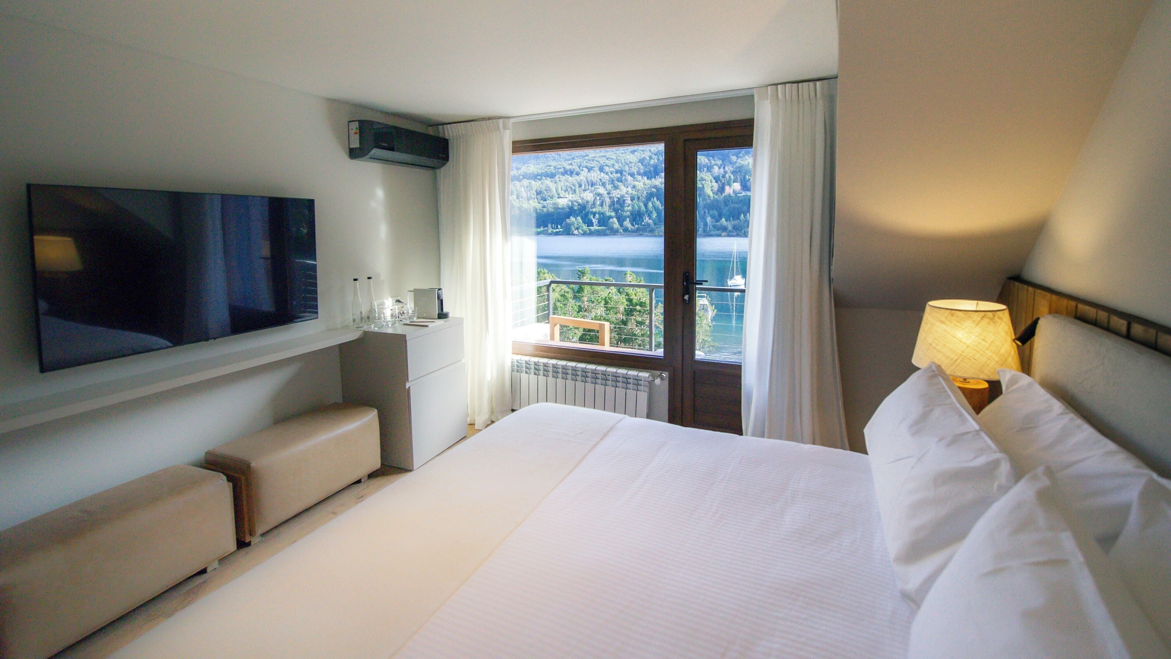 A hotel room with a bed, a TV, a desk with a chair, a window with a view of a lake, and a lamp.