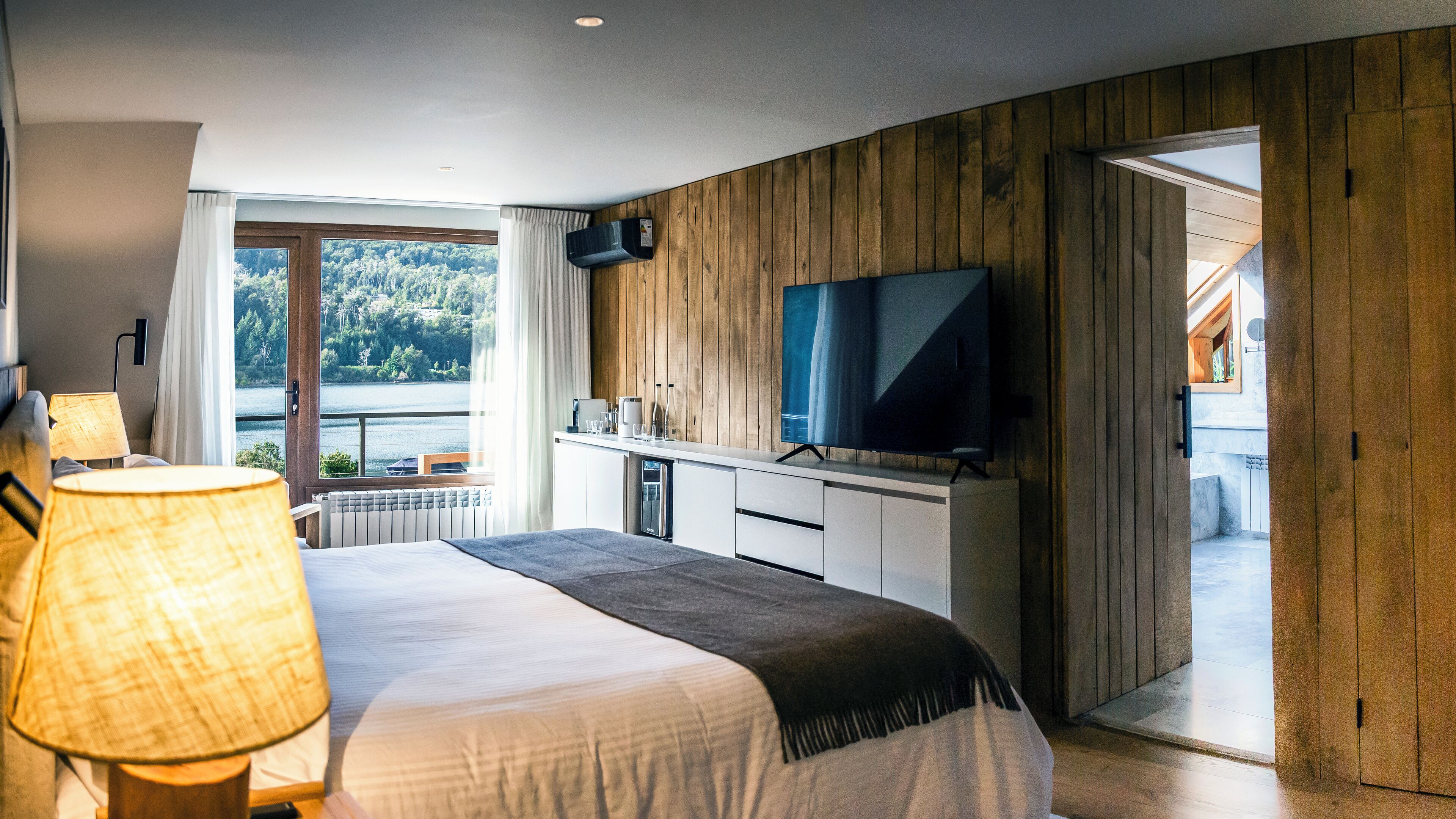 Une chambre d’hôtel moderne dotée d’un grand lit, d’un téléviseur à écran plat, d’un coin repas avec vue sur le lac et de lambris en bois sur les murs.