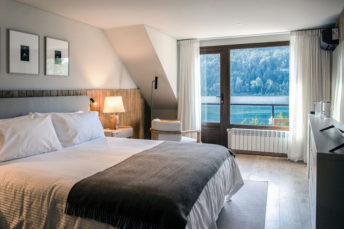 Ein modernes Hotelzimmer mit einem großen Bett, einem Nachttisch, einer Lampe, einem Sessel und Meerblick durch ein großes Fenster.