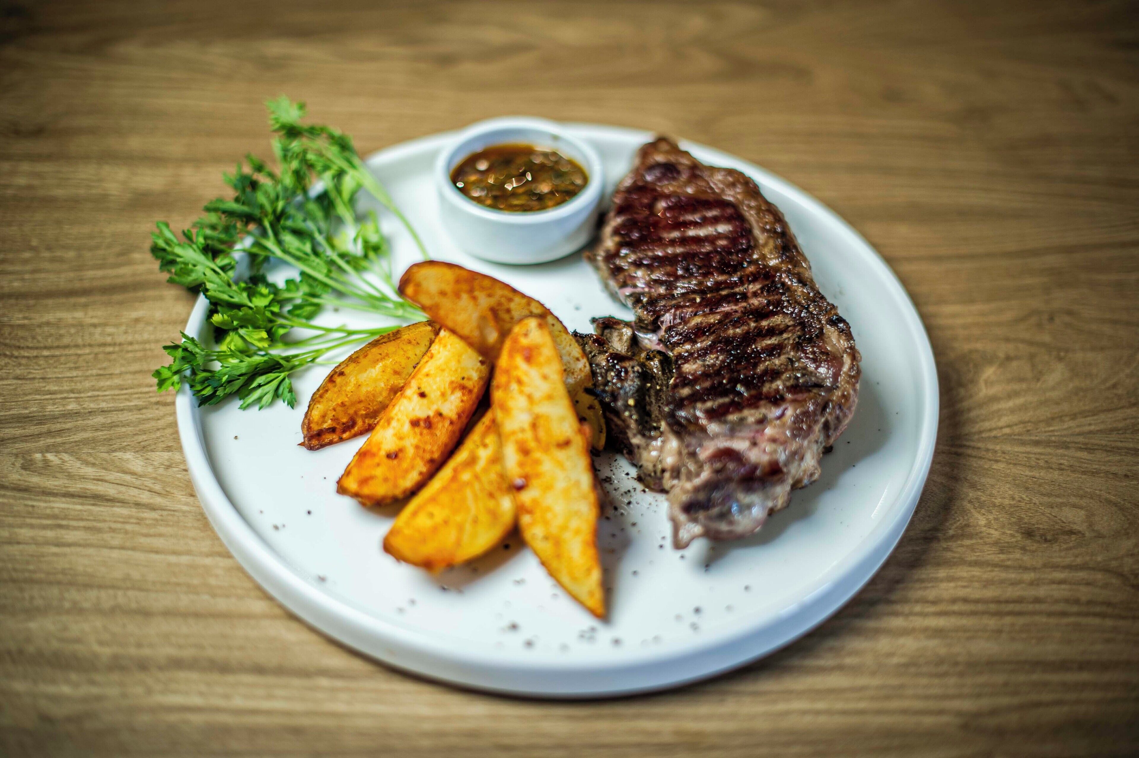 Un plato con un filete a la parrilla, patatas asadas y un recipiente pequeño con salsa, acompañado de una ramita de perejil, sobre una mesa de madera.