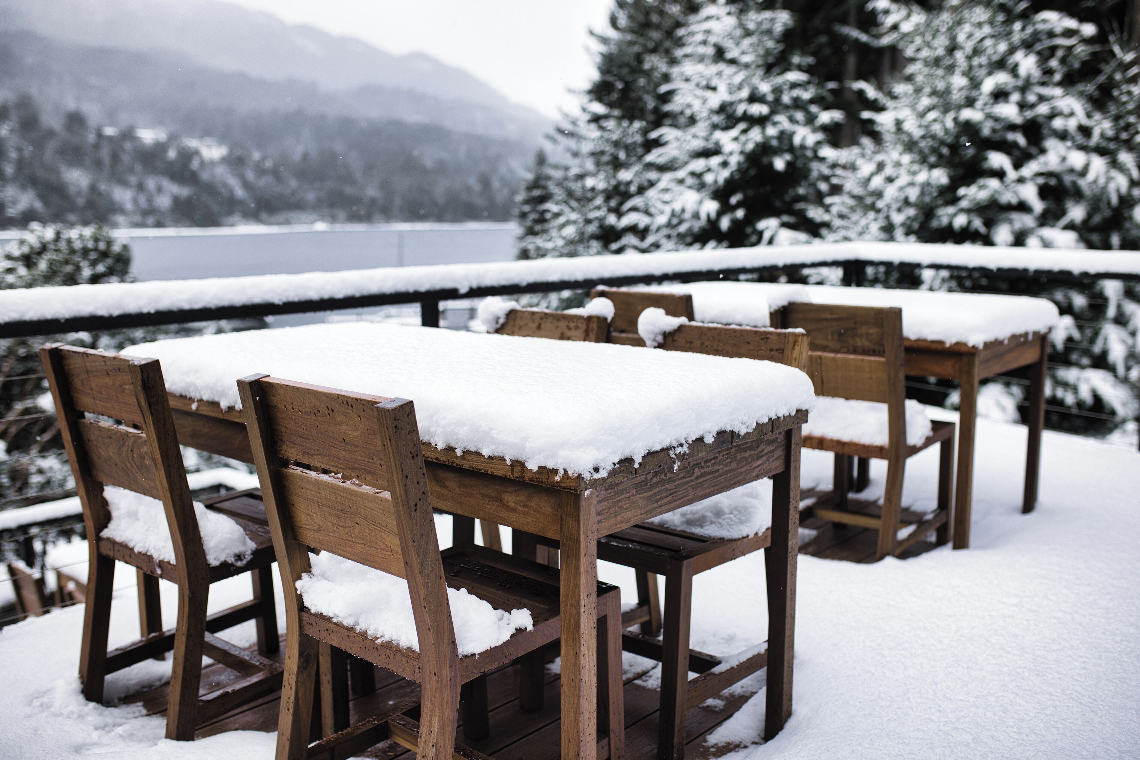 Outdoor wooden tables and chairs covered with snow, set against a snowy landscape with trees and a lake in the background.