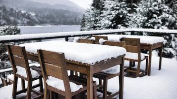 Outdoor wooden tables and chairs covered with snow, set against a snowy landscape with trees and a lake in the background.