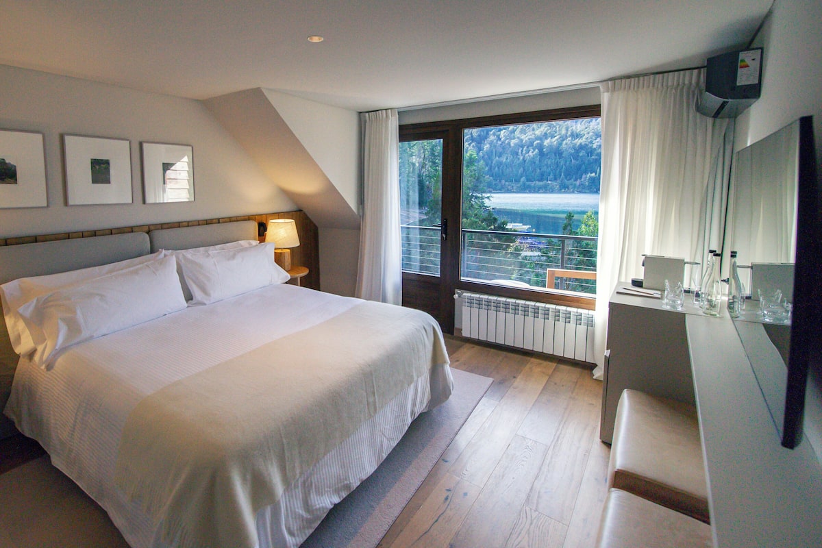 Ein Hotelzimmer mit einem großen Bett, zwei Nachttischlampen, Blick auf einen Balkon mit Geländer und einem an der Wand montierten Fernseher.