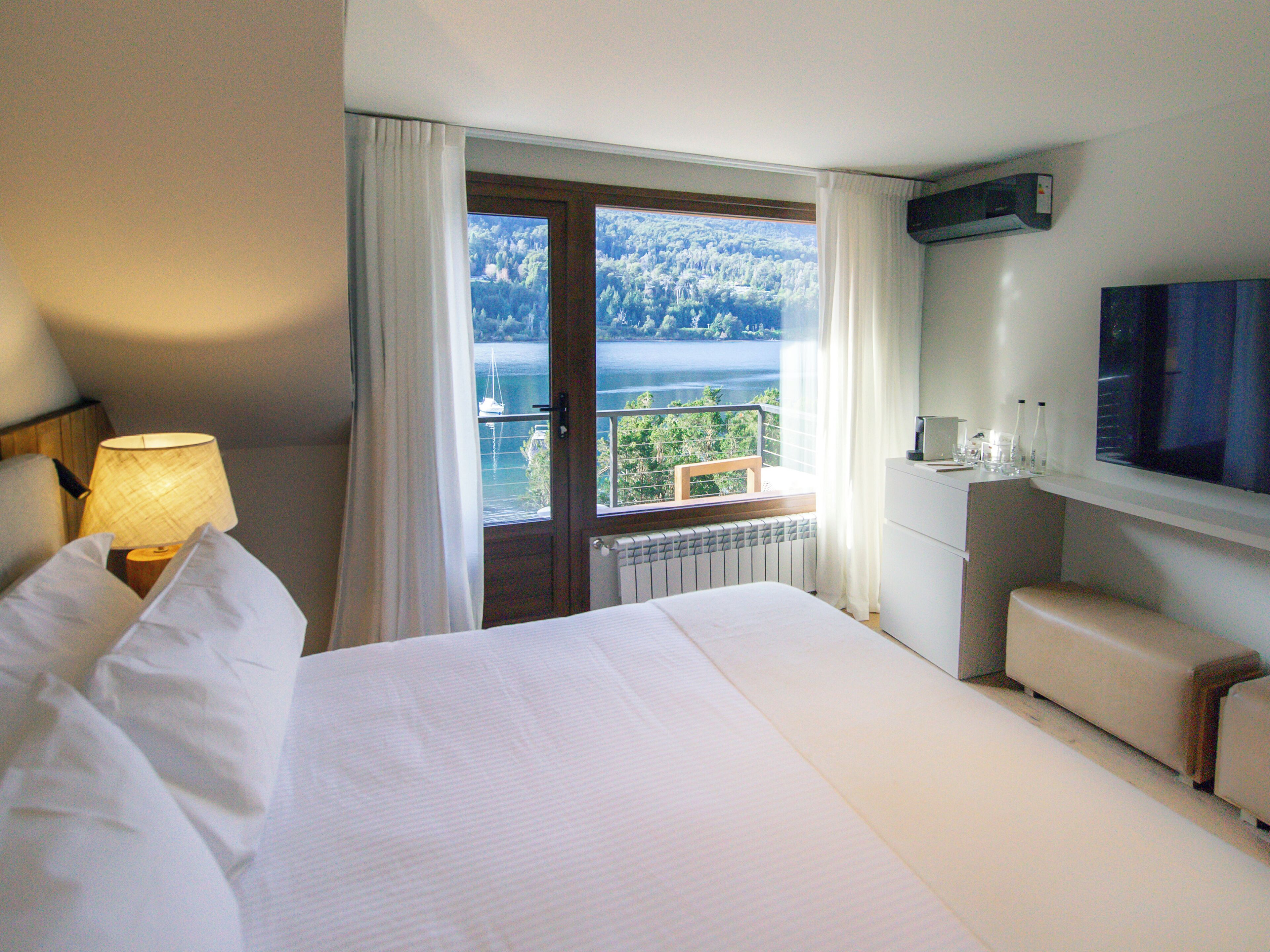 A hotel room with a bed, a desk, a chair, a TV, and a window with a view of a lake and trees.