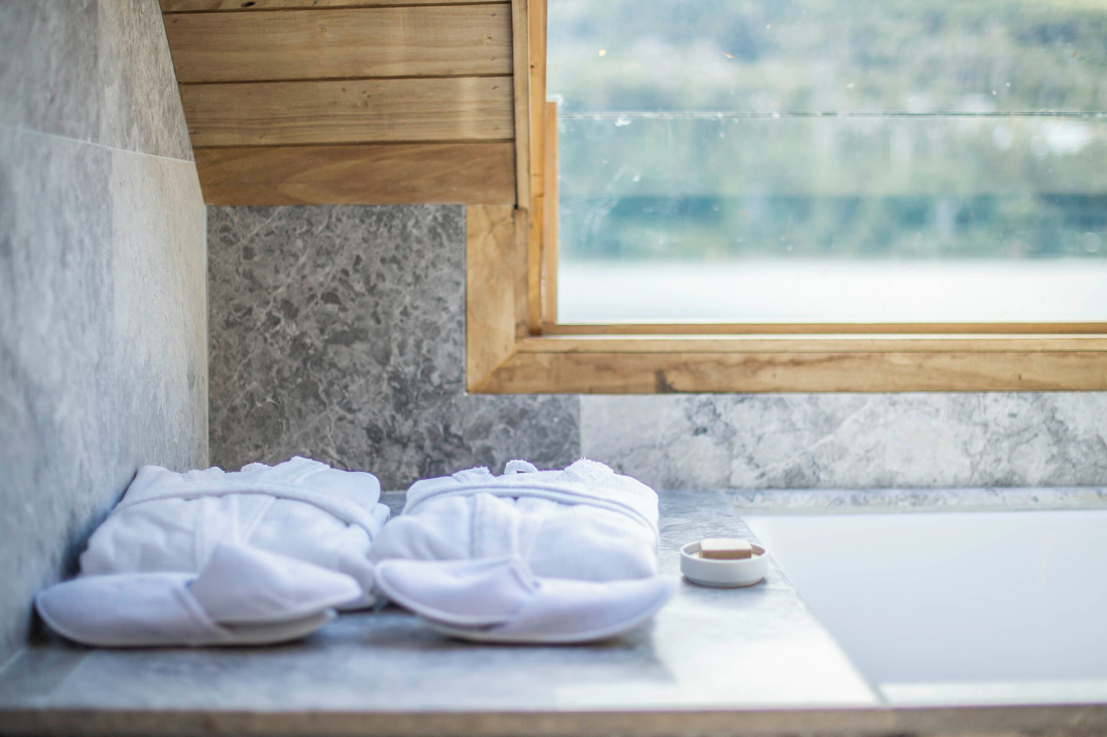 Un baño con techo de madera, pared de mármol y ventana. Hay dos toallas blancas y un jabonera en la encimera.