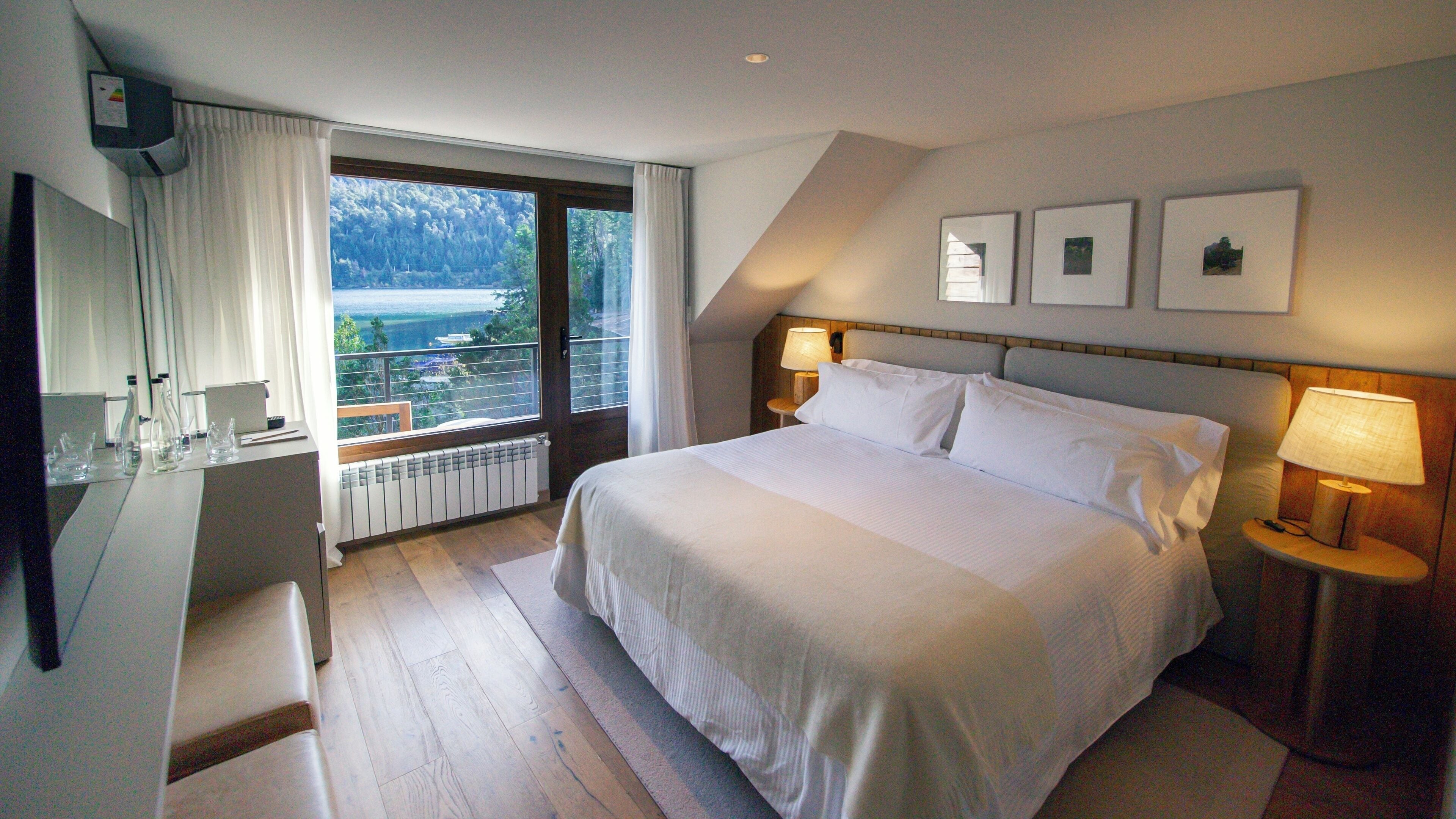A hotel room with a large bed, a desk with a computer, a window with a view of the outdoors, and two bedside lamps.