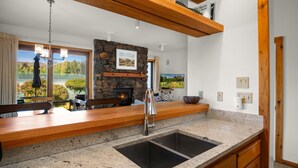 A modern kitchen with granite countertops, a stone fireplace, wooden beams, and a large window overlooking a scenic landscape.