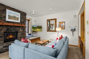 Una sala de estar con chimenea de piedra, un sofá gris, una mesa de centro de madera y un televisor.