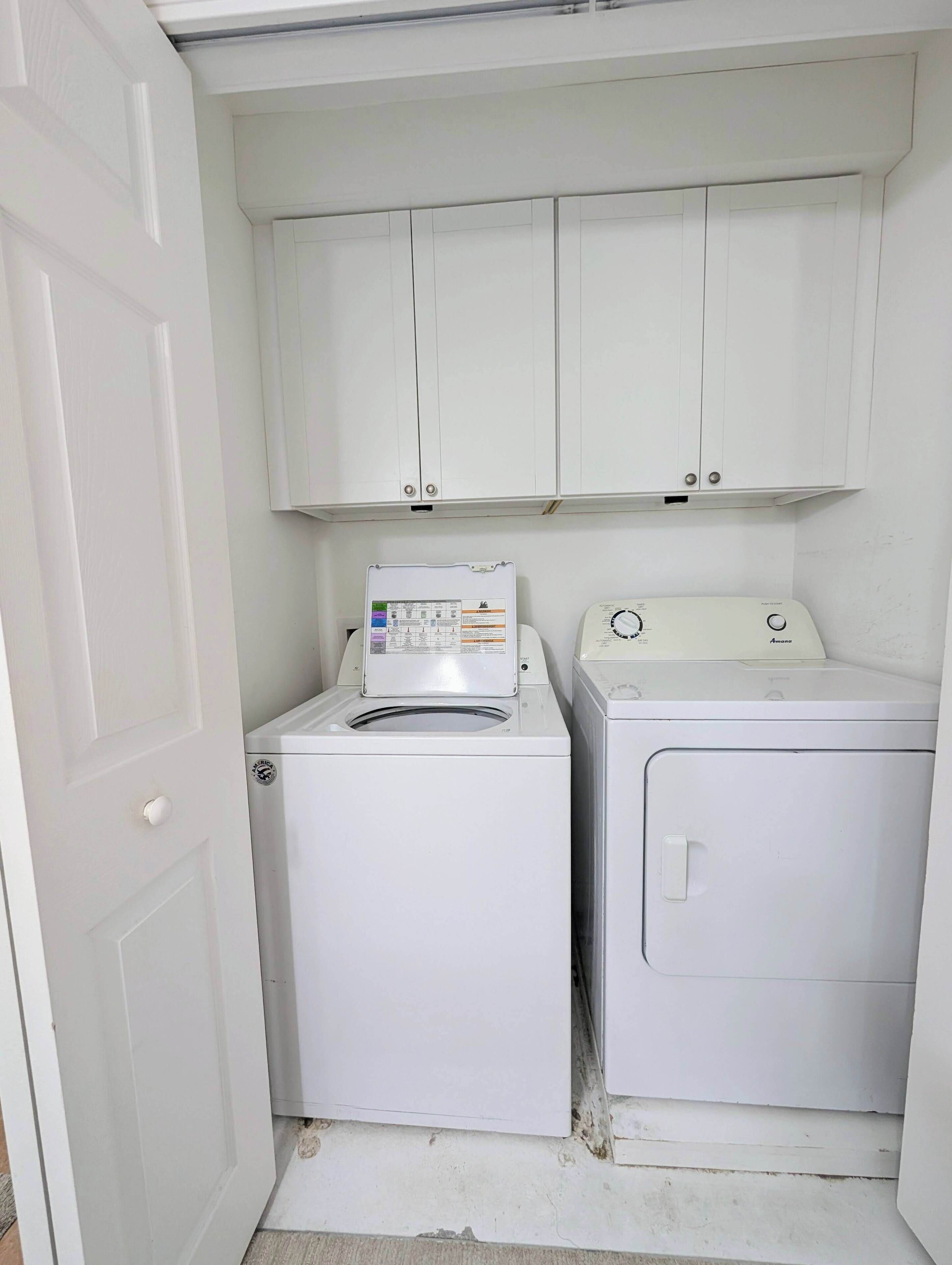 A laundry room with two white washing machines, a cabinet above, and a door on the left.