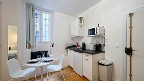 A compact kitchen with white cabinets, a microwave, and a coffee maker. There is a dining area with a white table and chairs, and a window with curtains.