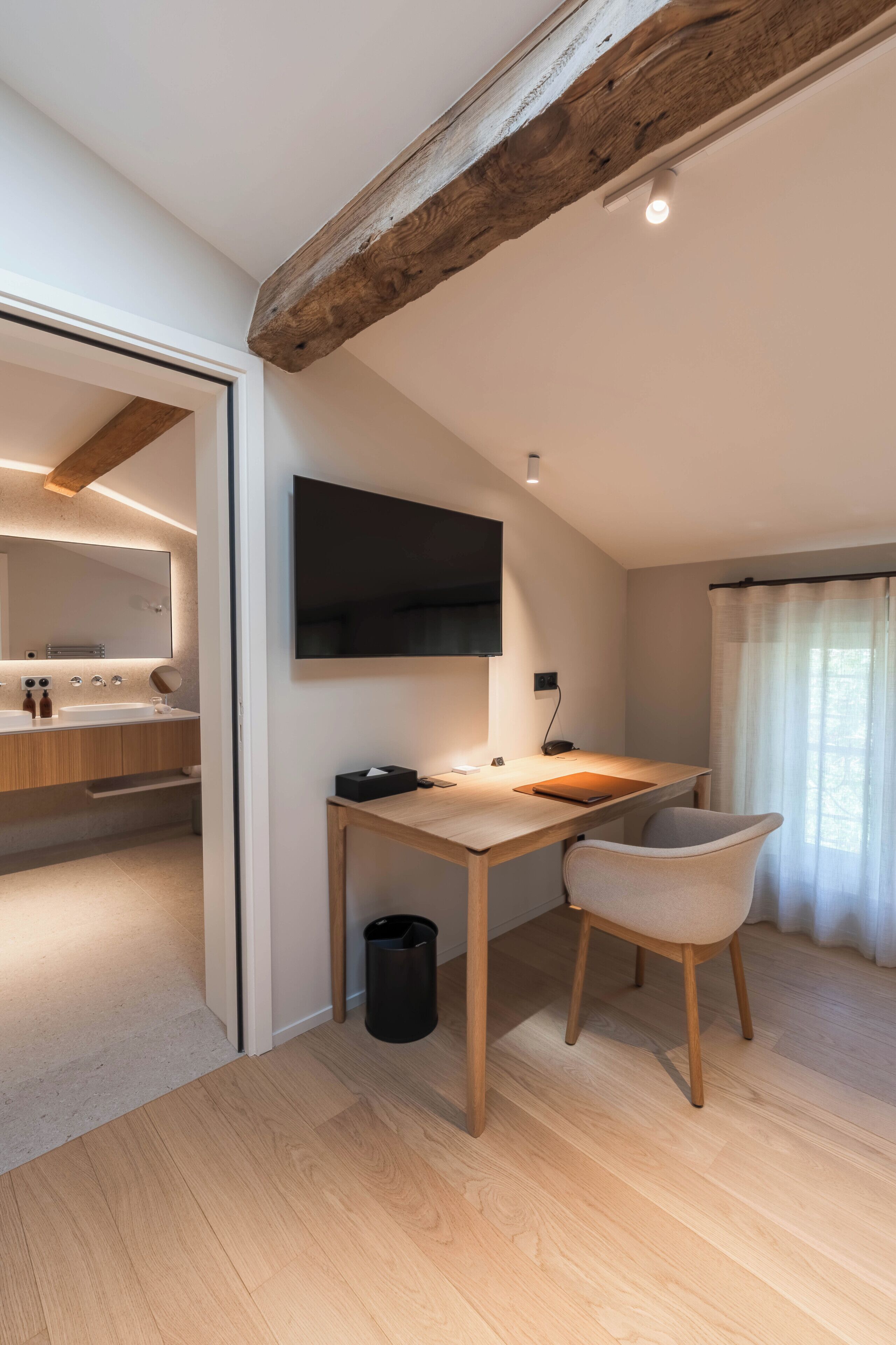 Una camera da letto moderna con soffitto in legno, una scrivania con sedia, una TV a schermo piatto e un bagno visibile attraverso una porta aperta.
