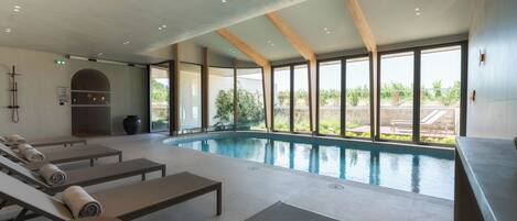 A modern indoor swimming pool with sun loungers and a view of the outdoors.