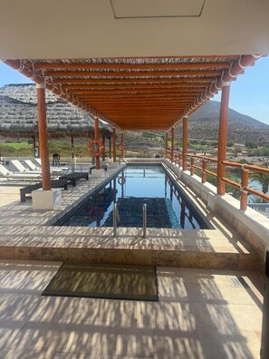 An outdoor swimming pool with a wooden pergola, lounge chairs, and a view of a mountainous landscape.