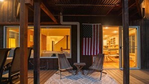 A wooden deck with two lounge chairs, an American flag, and a view into a kitchen area. - Riverfront Cabin Retreat – Unplug & Reconnect on the Entiat (Entiat)