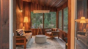 A cozy cabin interior with wooden walls, a large window offering a forest view, a sofa, a chair, and a round ottoman. - Riverfront Cabin Retreat – Unplug & Reconnect on the Entiat (Entiat)