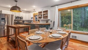 A modern kitchen with stainless steel appliances, a dining area with a wooden table set for four, and a large window offering a forest view. - Riverfront Cabin Retreat – Unplug & Reconnect on the Entiat (Entiat)