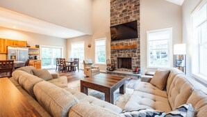 A spacious living room with a stone fireplace, a dining area, and a kitchen with wooden cabinets. - NF72: Brand New, Creekside home, 3 King Suites, Hot Tub. Firepit (Davis)