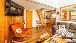A cozy living room with a wooden floor, a leather sofa, a rocking chair, and a television mounted on the wall. - Cozy Updated Cabin, close to towns, A/C (Davis)