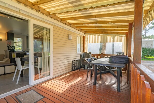 A covered patio with wooden flooring, a table with chairs, and a sliding glass door leading to an indoor area with a kitchen.
