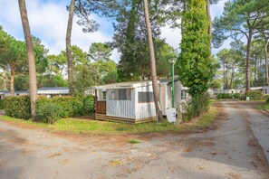A small, single-story mobile home with a porch, surrounded by trees and a clear sky.