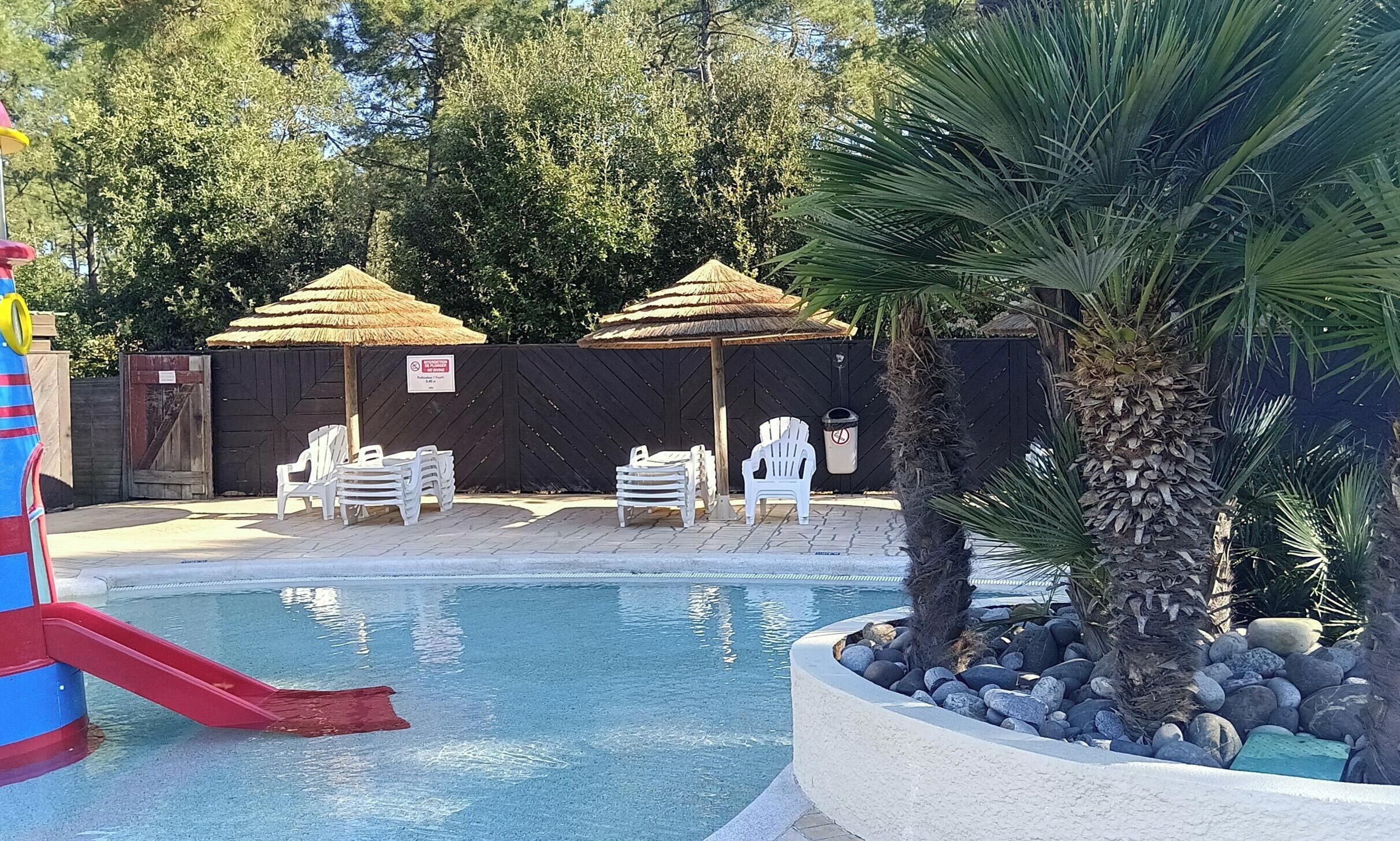 En swimmingpool med solstolar och parasoller av halmtak, omgiven av träd och ett staket av trä.