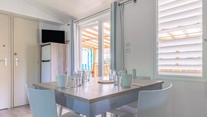 A dining area with a table set for four, a television above the table, and a view of the outside through large windows. - Mobile Home on Oléron in 5-Star Campsite with Shared Pool (Saint George d'Oleron)