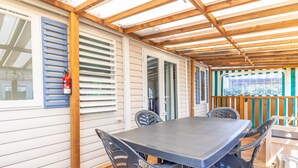 A covered patio with a table and chairs, wooden beams, a fire extinguisher, and a window with blinds. - Mobile Home on Oléron in 5-Star Campsite with Shared Pool (Saint George d'Oleron)