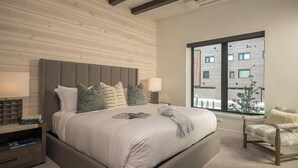 Une chambre moderne avec un grand lit, une table de chevet avec des livres, une chaise et une fenêtre donnant sur des immeubles.