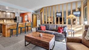 Ein modernes Wohnzimmer mit einer Couch, einem Couchtisch und Sesseln. Es gibt eine Küchenzeile mit einem Kühlschrank und einem Esstisch.