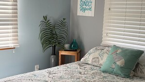 Una camera da letto con un letto, una ventola a soffitto, una lampada a parete, una pianta in vaso, un tavolino con un vaso e un telecomando, e un cartello a parete con del testo.