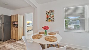 Zona de menjador amb una taula rodona, cadires blanques i un vas amb flors vermelles. Hi ha una cuina amb un frigorífic i armaris de fusta. Al mur hi ha un quadre, i es veu una finestra amb persianes.