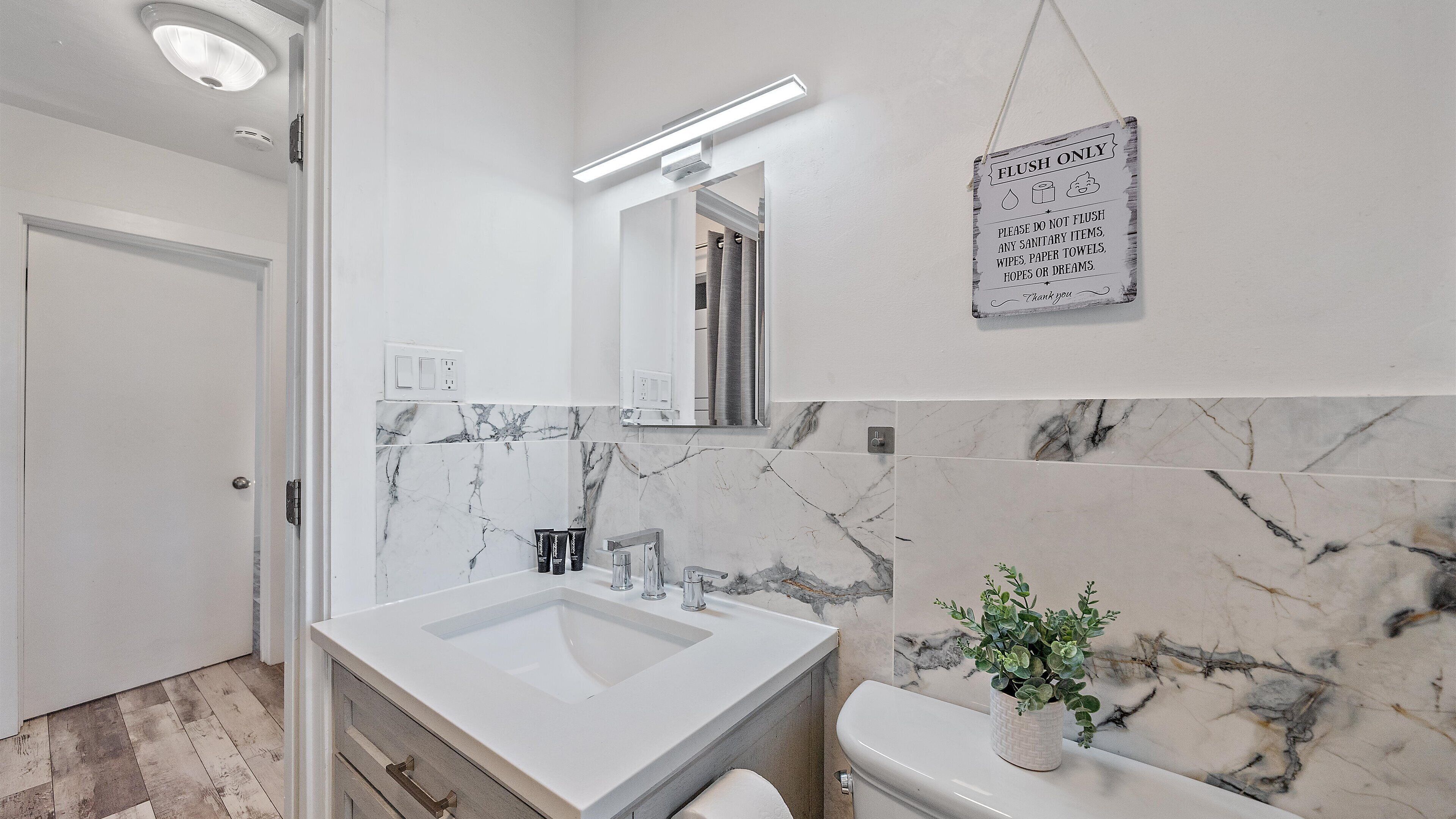 Une salle de bain avec un lavabo blanc, un miroir et des toilettes. Un panneau est affiché sur le mur avec les inscriptions suivantes : « NE PAS JETER QUE LES DÉCHETS » et « VEUILLEZ NE PAS JETER DANS LES TOILETTES : NI ARTICLES D’HYGIÈNE, NI LINGETTES, NI ESSUIE-TOUT, NI ESPOIRS, NI RÊVES. »