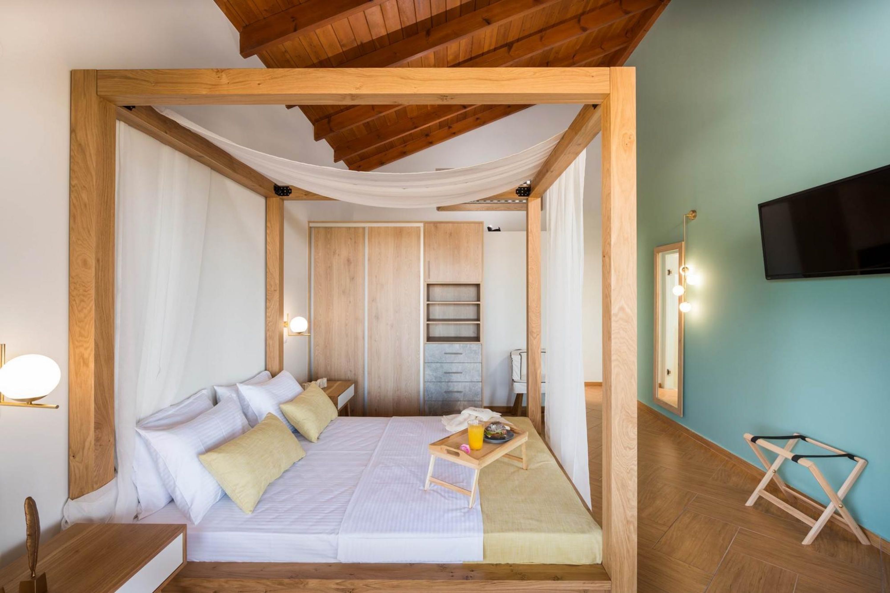 A bedroom with a wooden frame, a bed with pillows, a bedside table, a tray with a drink, and a TV on the wall.