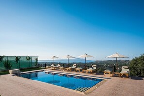 A pool area with lounge chairs and umbrellas, surrounded by a fence and a view of the landscape. - Erato Villa in Gavalochori (Apokoronas)