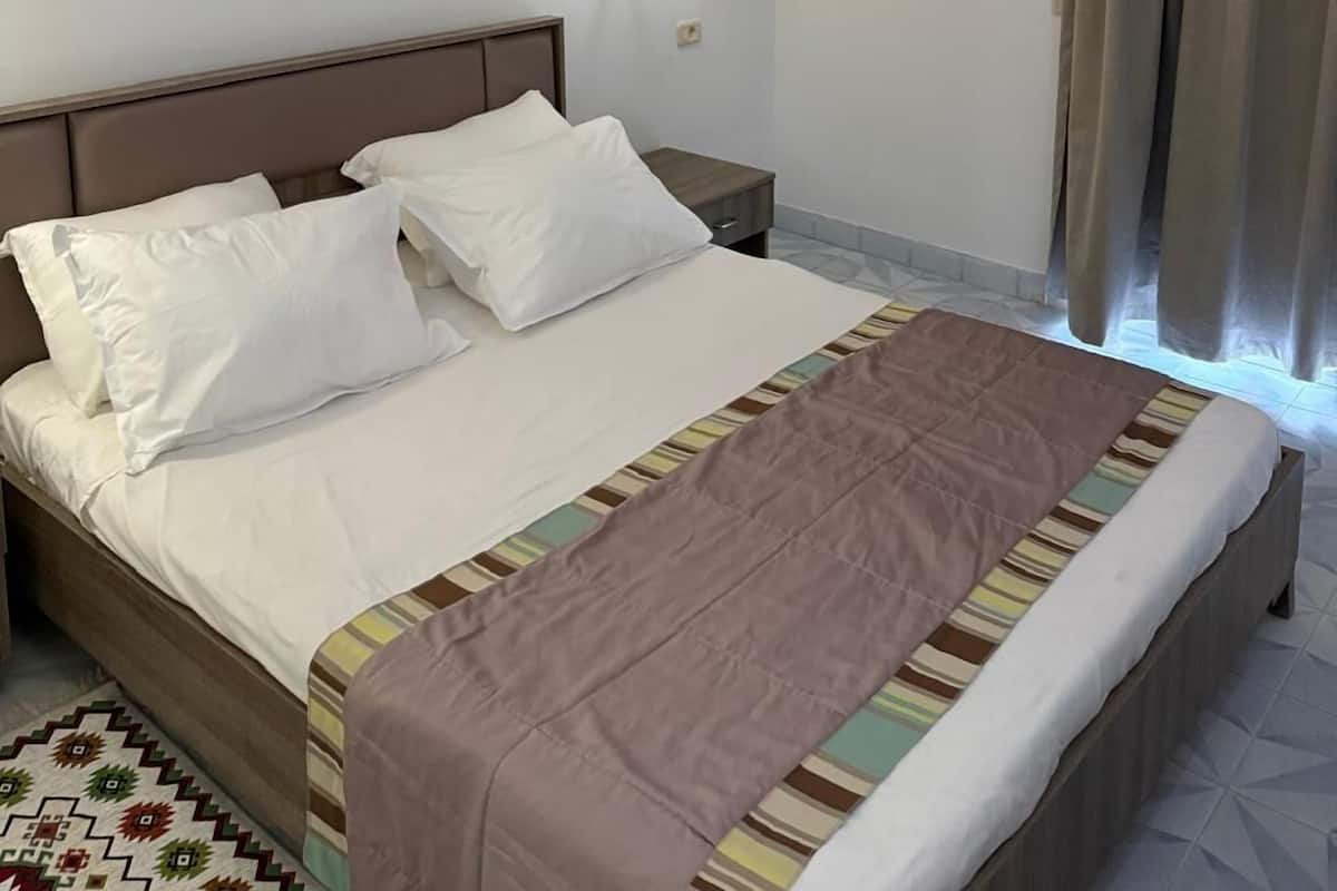 A hotel room with a bed, a nightstand, a wall-mounted lamp, a painting, and a window with curtains.