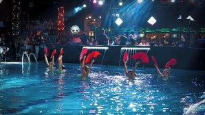 A swimming pool with people performing synchronized routines, a stage with a DJ, and a crowd in the background.
