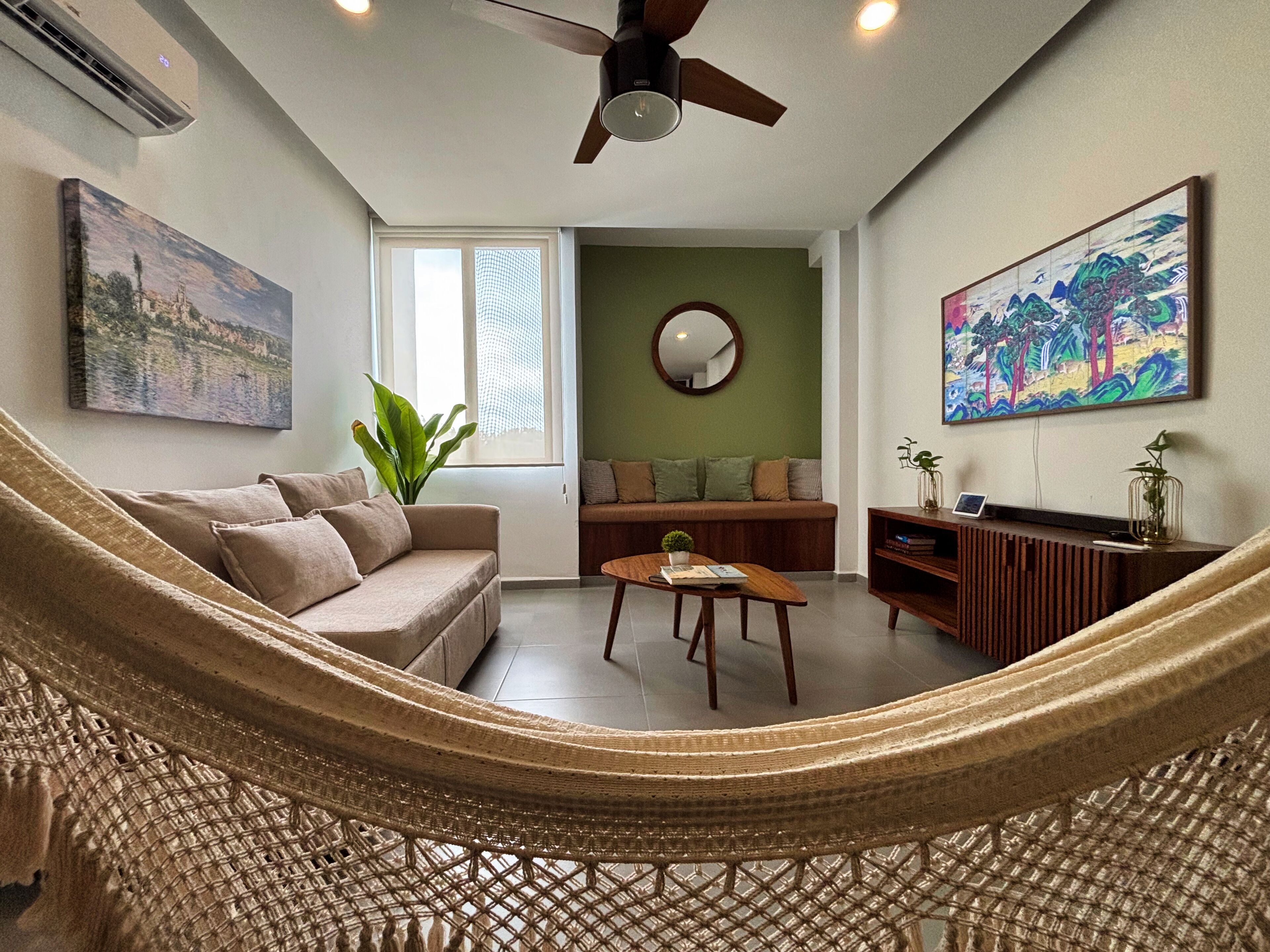 A living room with a hammock, a sofa, a coffee table, a TV stand, and artwork on the walls.