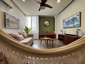 A living room with a hammock, a sofa, a coffee table, a TV stand, and artwork on the walls. - Loft with Pool and King Size Bed in Montejo (Mérida)