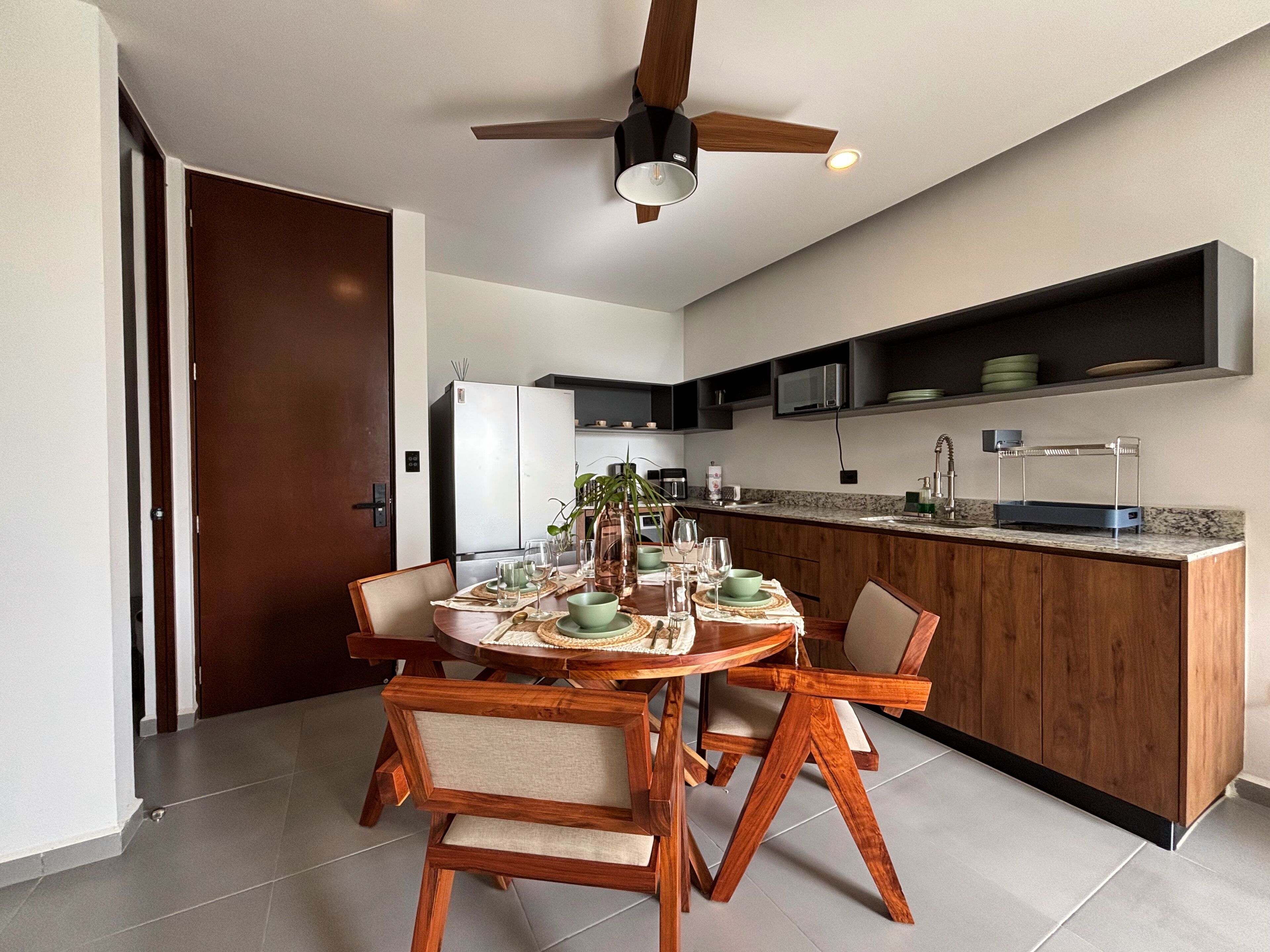 A modern kitchen with a dining area, featuring a round wooden table, chairs, a ceiling fan, and built-in appliances.