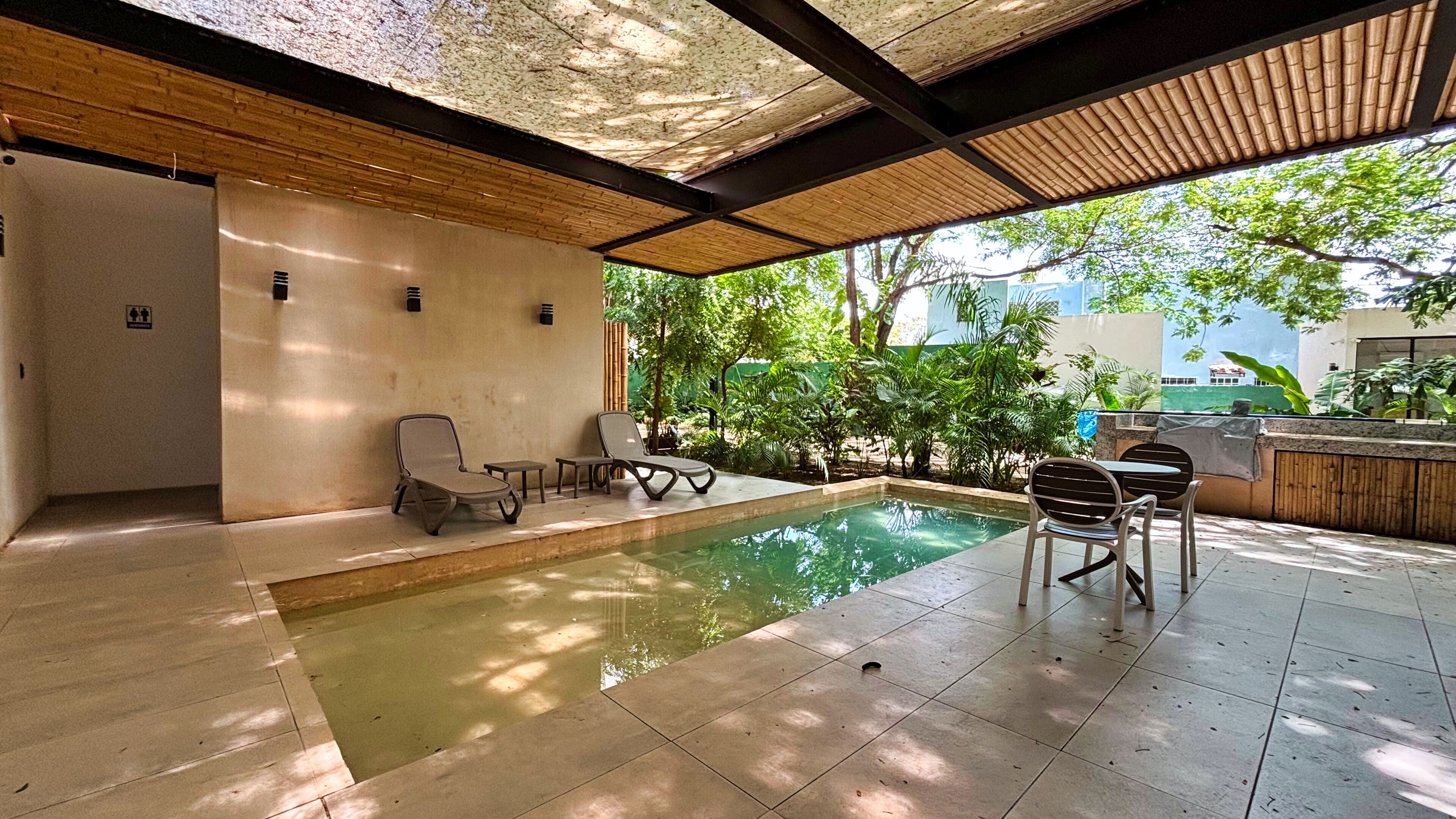 A pool area with lounge chairs and a table, surrounded by greenery and a covered patio.