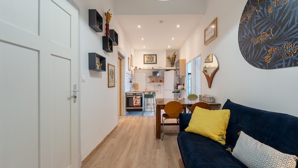 Een modern interieur met een houten vloer, een donkerblauwe bank met gele en gemêleerde kussens, een eethoek met een houten tafel en stoelen, en een keuken met witte apparaten.