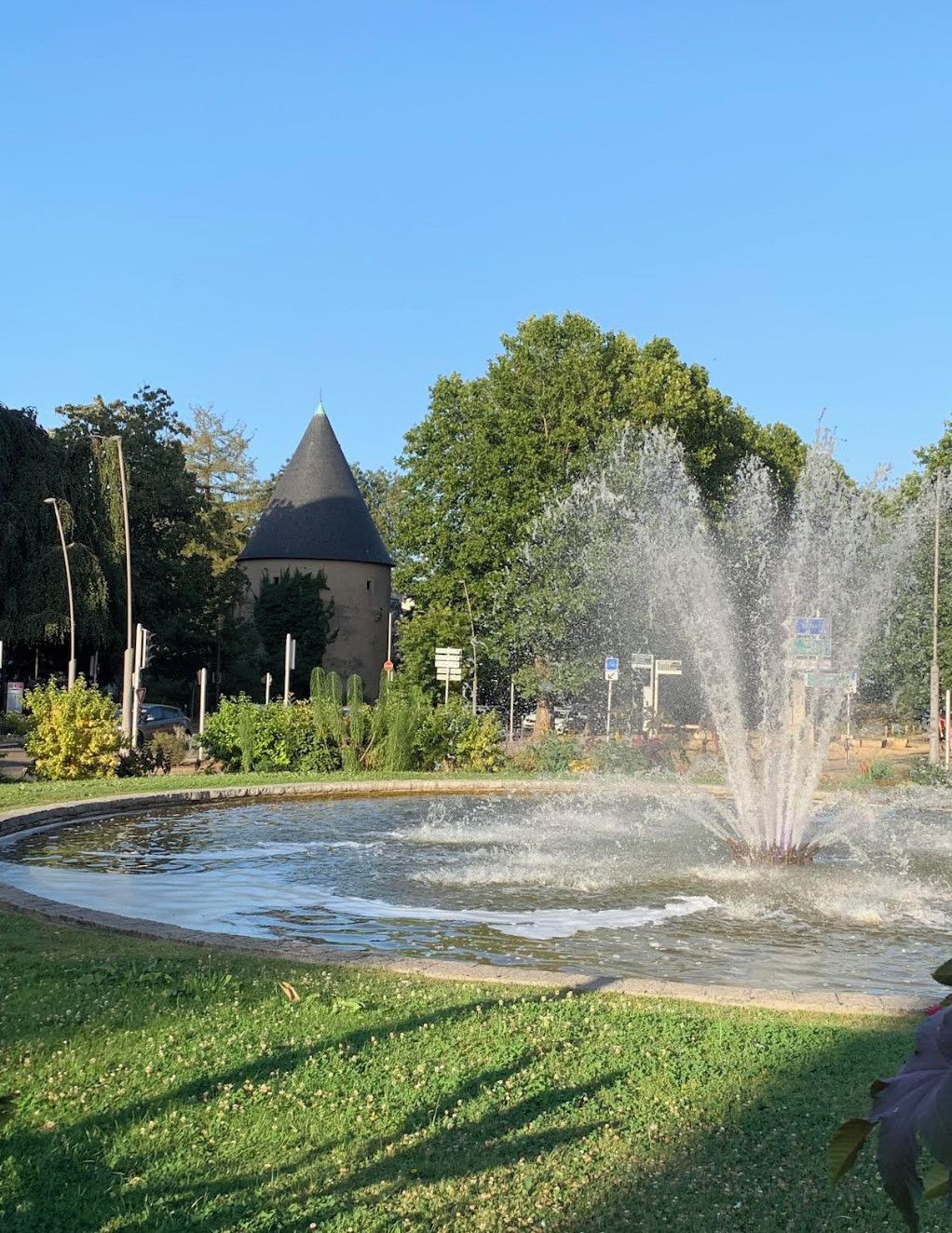 A fountain with water jets, a grassy area, and a building with a conical roof.