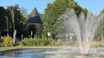 Une fontaine avec des jets d’eau, une pelouse et un bâtiment surmonté d’un toit conique.