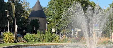 Una fuente con chorros de agua, un área con césped y un edificio de techo cónico.