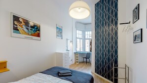Een slaapkamer met een bed, een bureau en een stoel. Er zijn twee ramen en een deur.