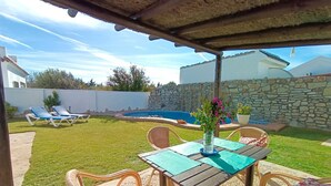 A covered patio with a table and chairs, a swimming pool, and a stone wall in the background.